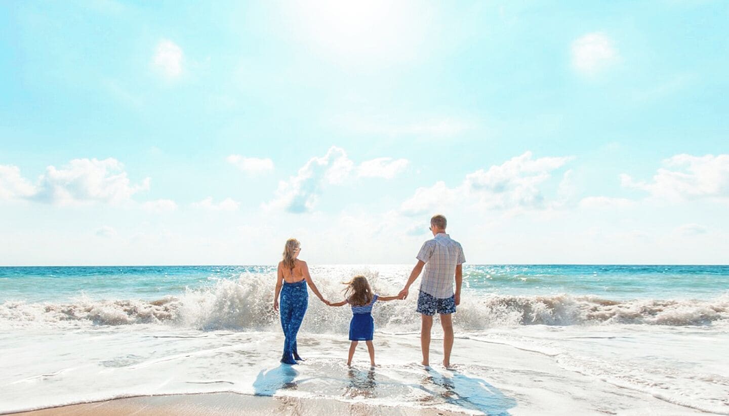 Eine dreiköpfige Familie hält auf einem Strand Händchen, während Wellen rollen.