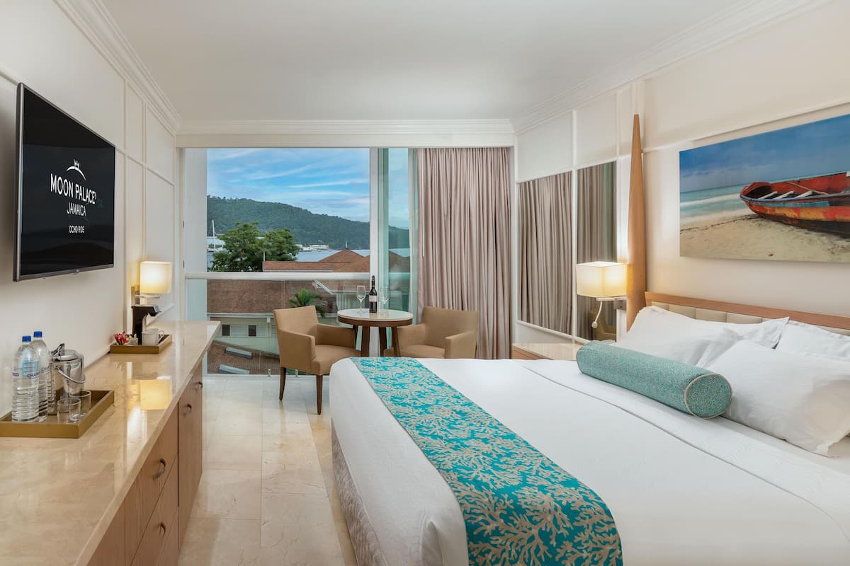 A hotel room with a bed, a desk with a computer, a chair, a small table with a bottle and glasses, and a view of the beach and mountains.
