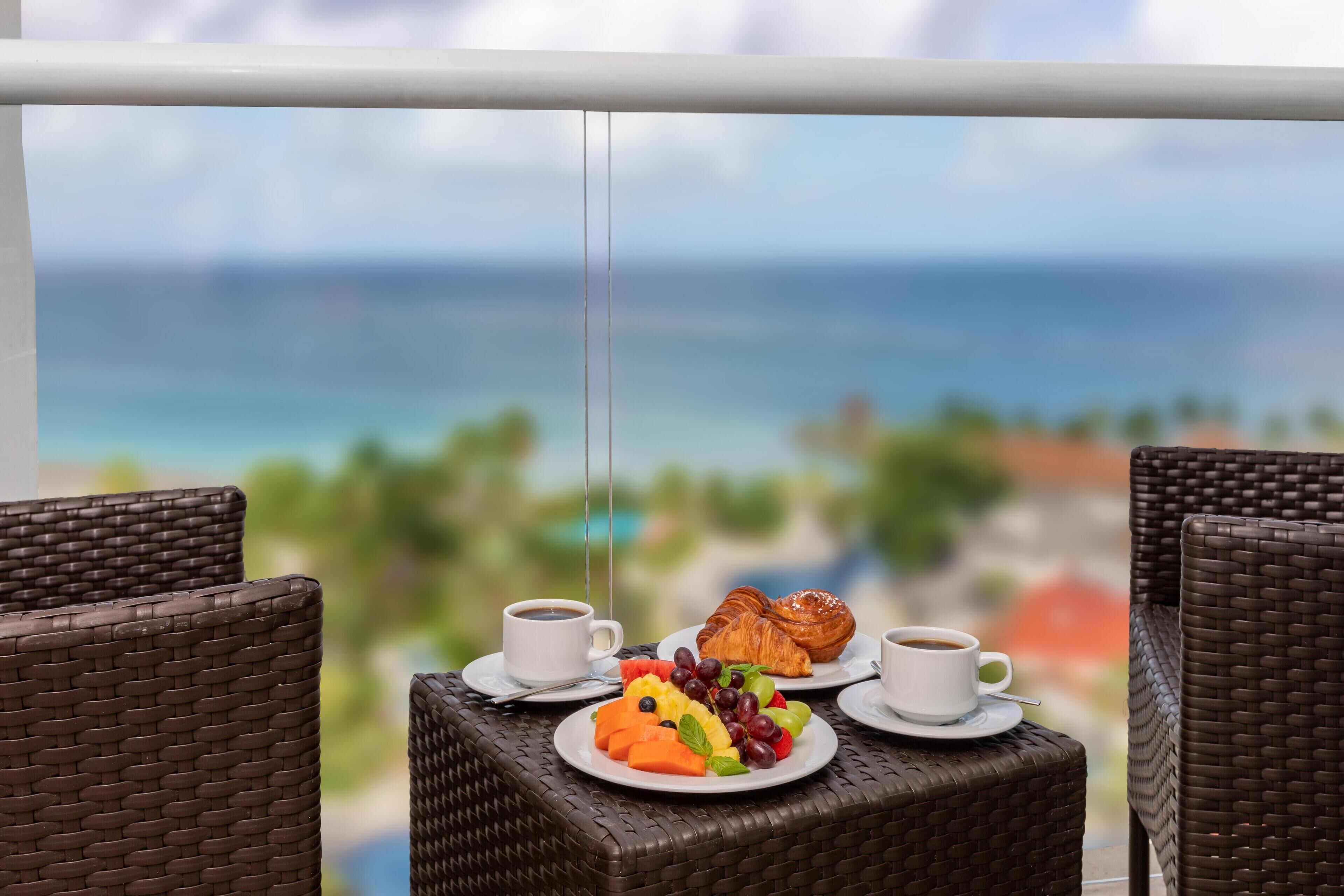 A wicker table with a croissant, fruit, and coffee cups, overlooking a beach.