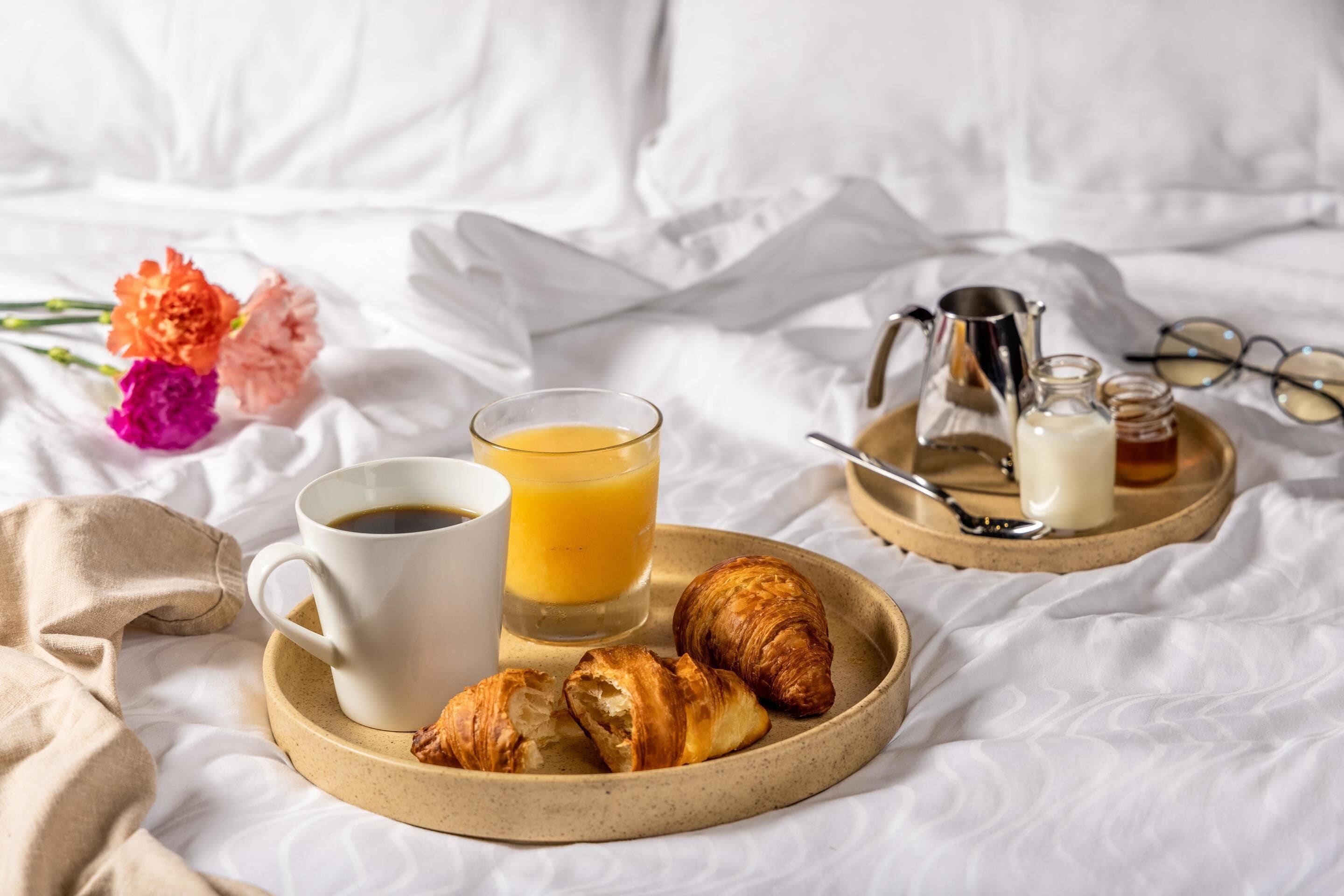 Ein Frühstücksservice auf einem Bett mit einer Kaffeetasse, einem Glas Orangensaft, Croissants, einer Schale mit Milch und einer Marmeladenglas.