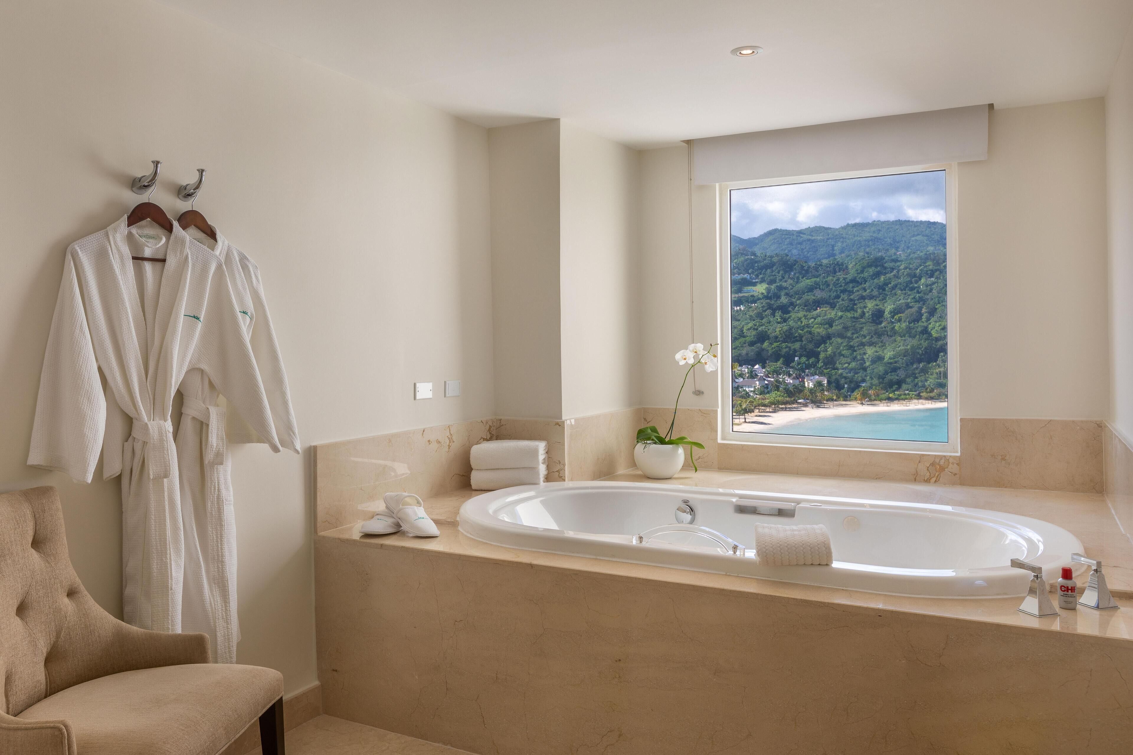 A bathroom with a large bathtub, a view of a beach and mountains, and a robe hanging on the wall.