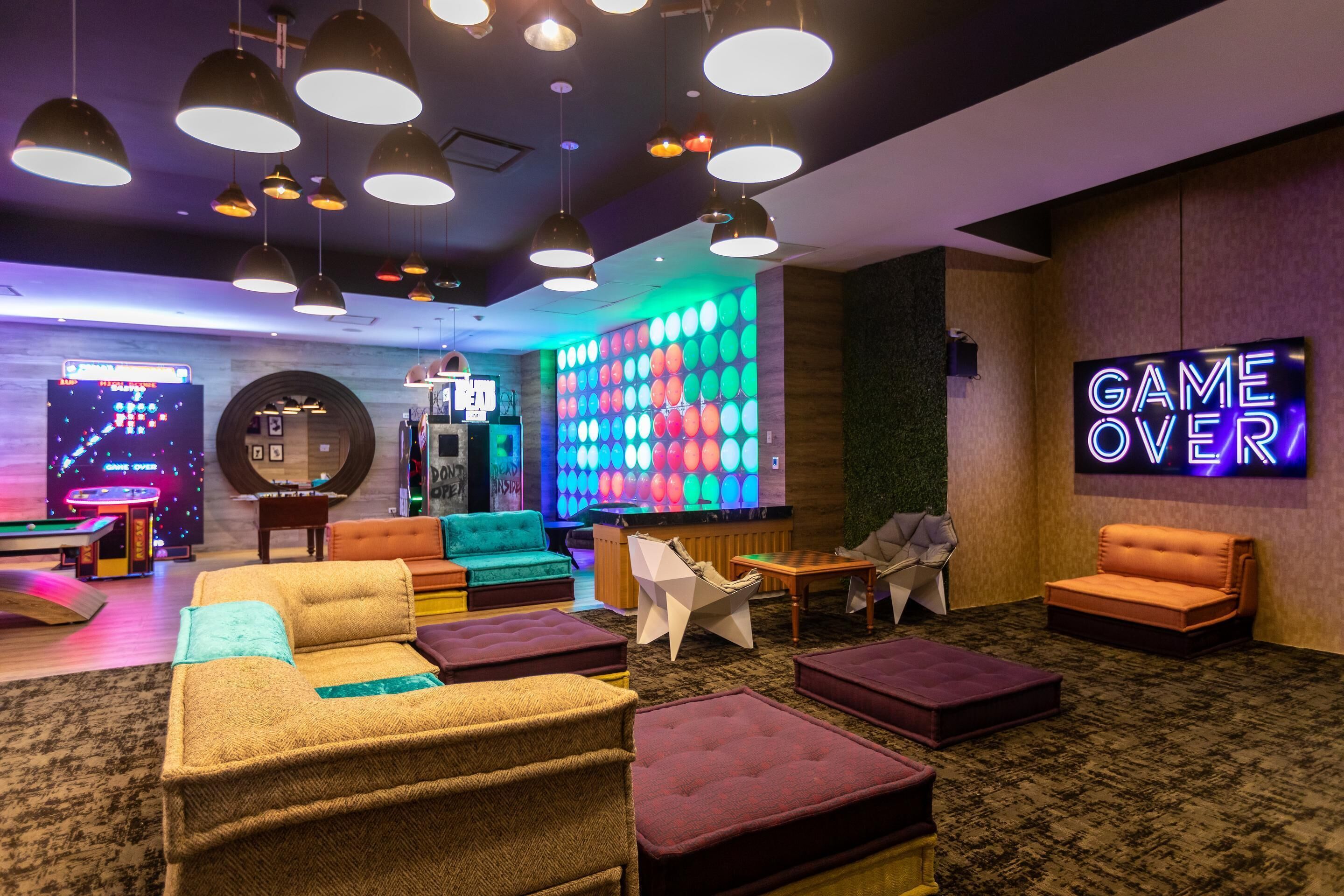 Un espace détente avec différents types de sièges, notamment des canapés et des poufs, un panneau néon indiquant « PARTIE TERMINÉE » et un mur décoré de motifs colorés.