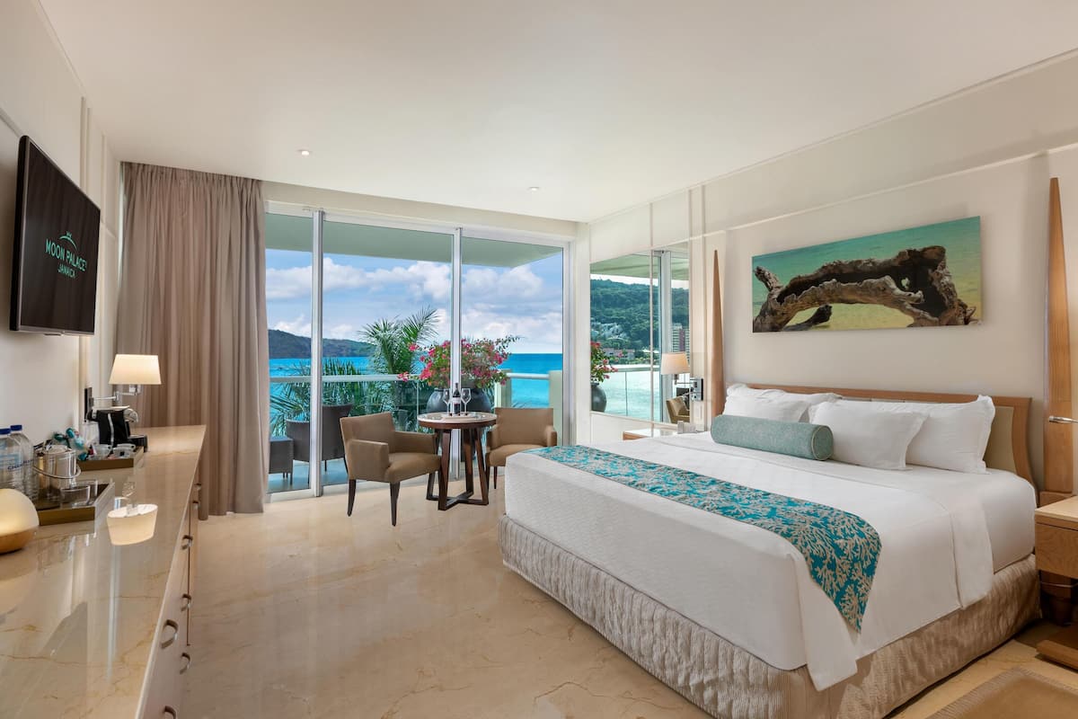 A hotel room with a large bed, a TV, a balcony with a view of the sea, and a dining area.