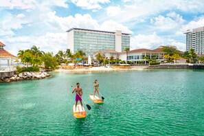 A resort with a large building, a beach, and two people paddleboarding.