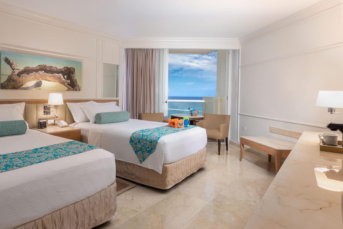 A hotel room with two beds, a desk, a chair, and a view of the ocean.