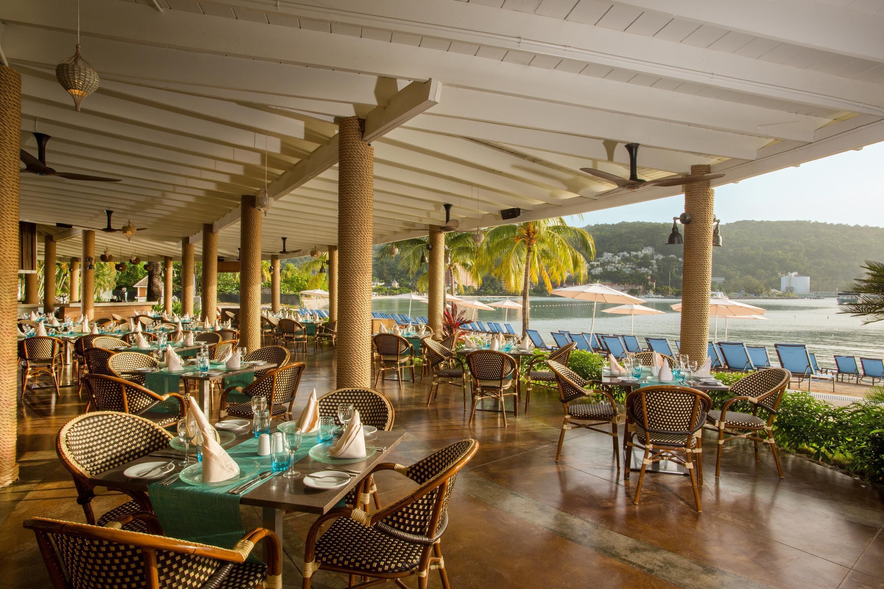 A covered outdoor dining area with wicker furniture and tables set for dining, overlooking a beach and palm trees.