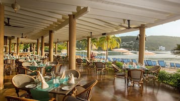 A covered outdoor dining area with wicker furniture and tables set for dining, overlooking a beach and palm trees.