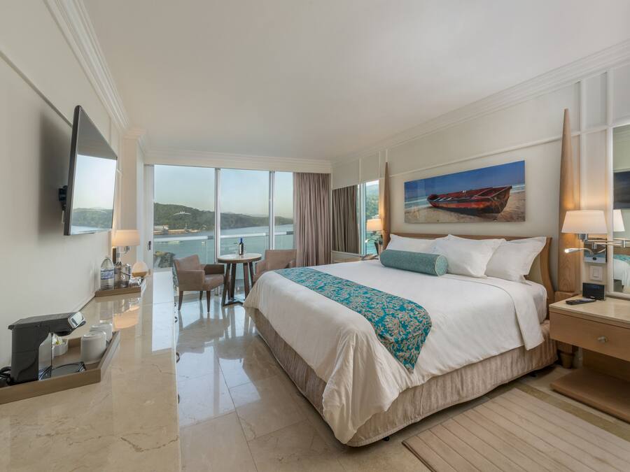 A hotel room with a large bed, a desk, a chair, a TV, and a painting of a boat on the wall.
