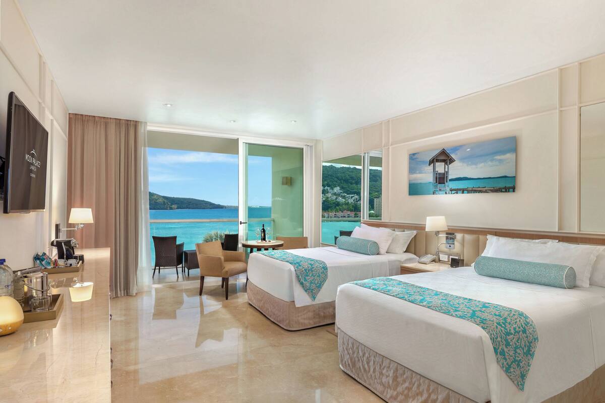 A hotel room with two beds, a TV, a desk with a lamp, and a view of the sea.