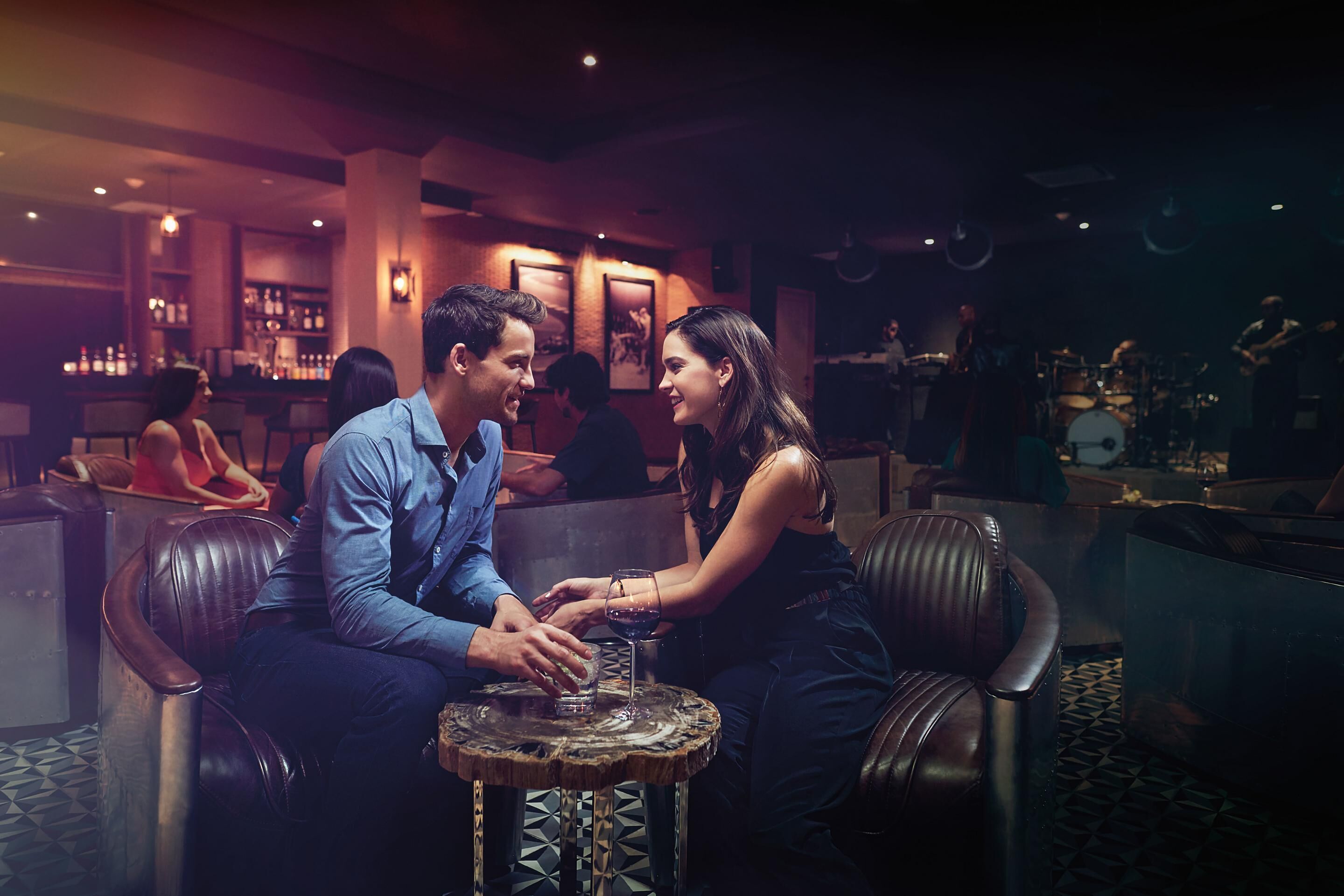 A couple seated at a round table in a dimly lit bar area with leather seating and a bar in the background.