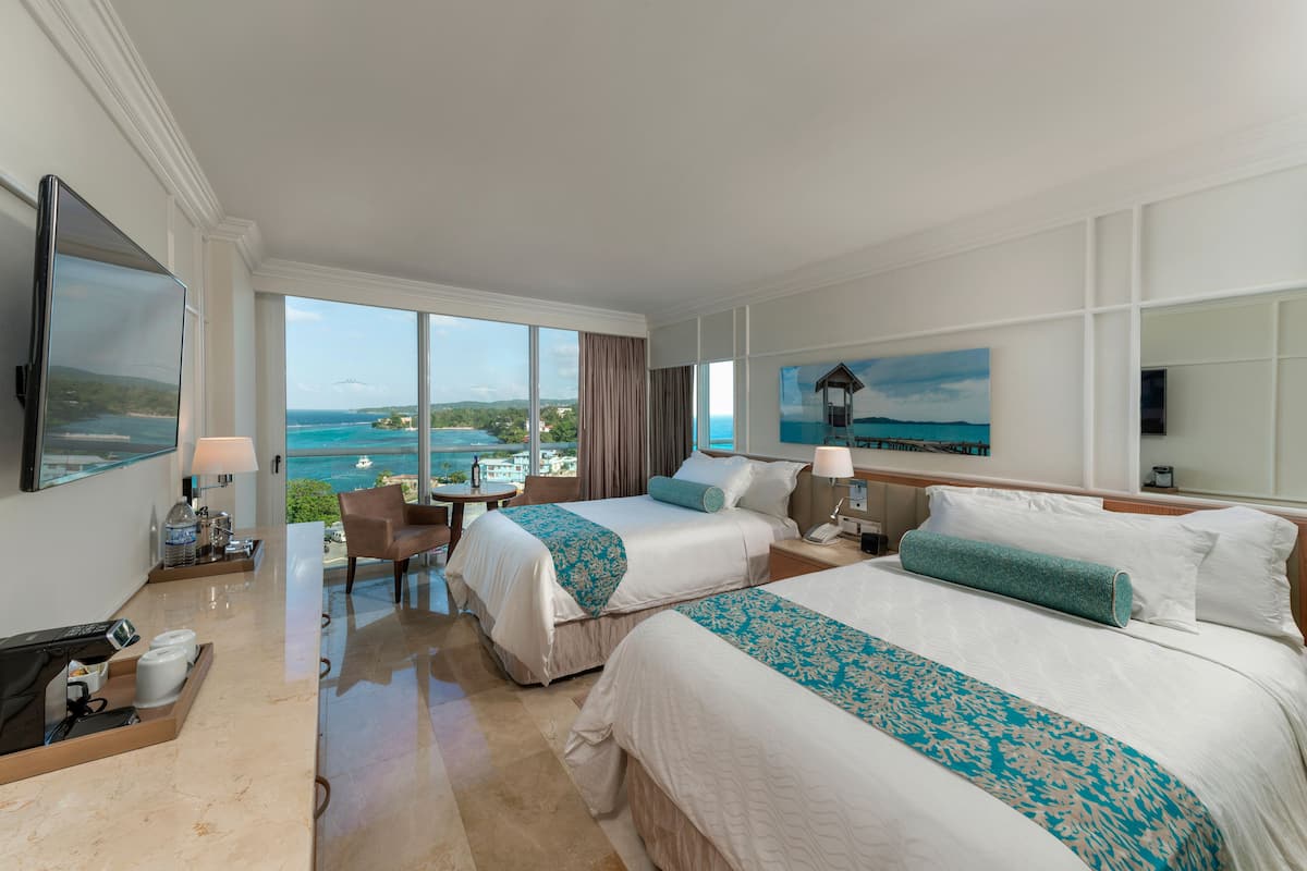 A hotel room with two beds, a desk, a chair, and a large window with a view of the sea.
