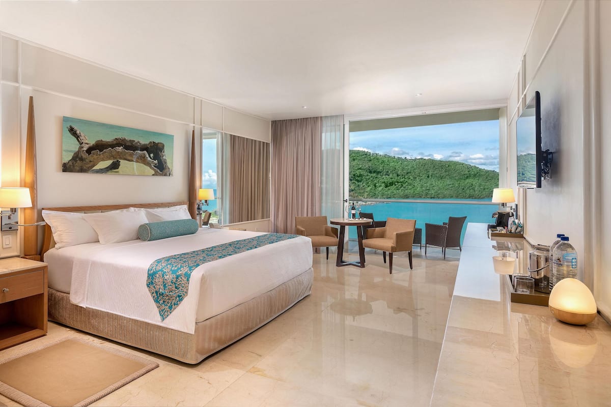 A hotel room with a large bed, a desk, a chair, and a view of a green landscape.