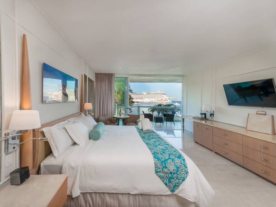 A modern hotel room with a large bed, a TV, and a balcony with a view of a cruise ship.