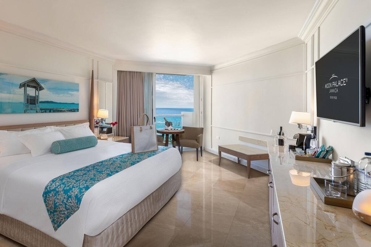 A hotel room with a large bed, a desk, a chair, a TV, and a view of the ocean.