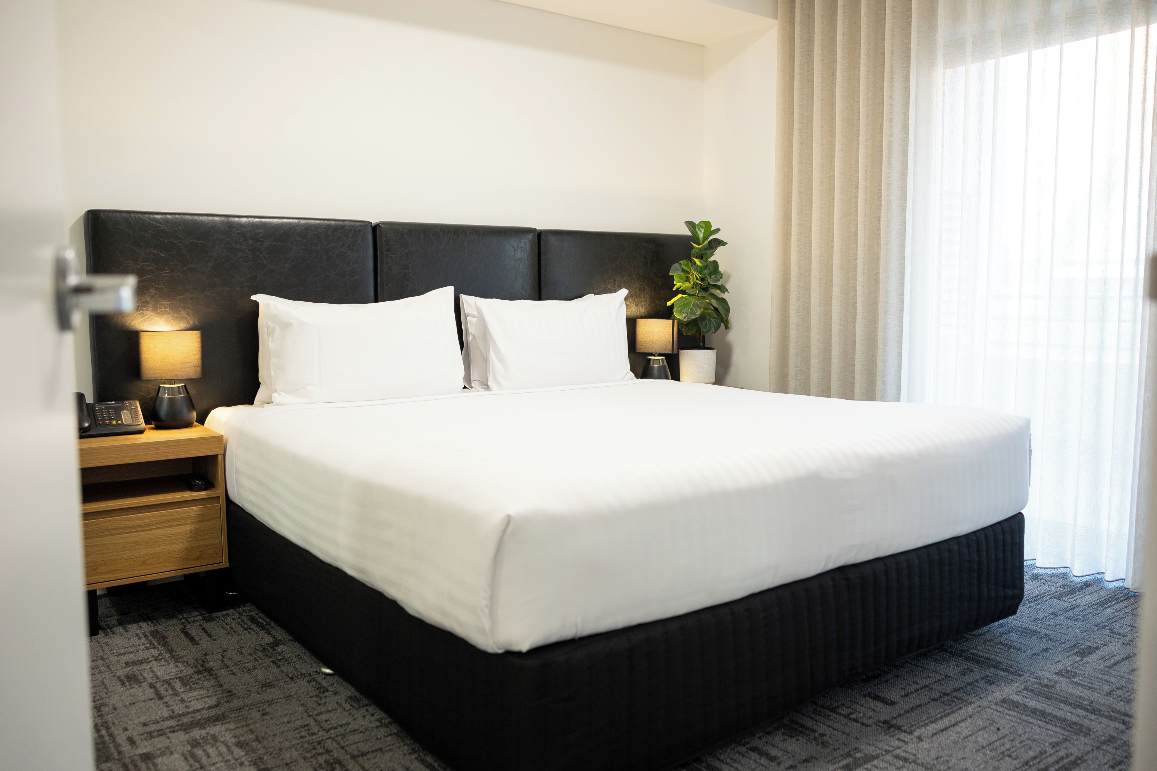 A hotel room with a large bed, two bedside tables, a lamp, a plant, and a window with curtains.