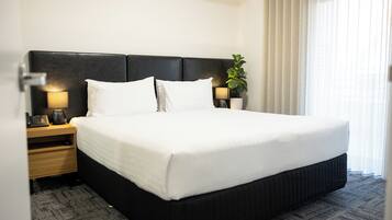 A hotel room with a large bed, two bedside tables, a lamp, a plant, and a window with curtains.
