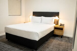 A neatly made bed with white linens, a dark headboard, two bedside lamps, a wooden nightstand with a phone, and a patterned carpet.