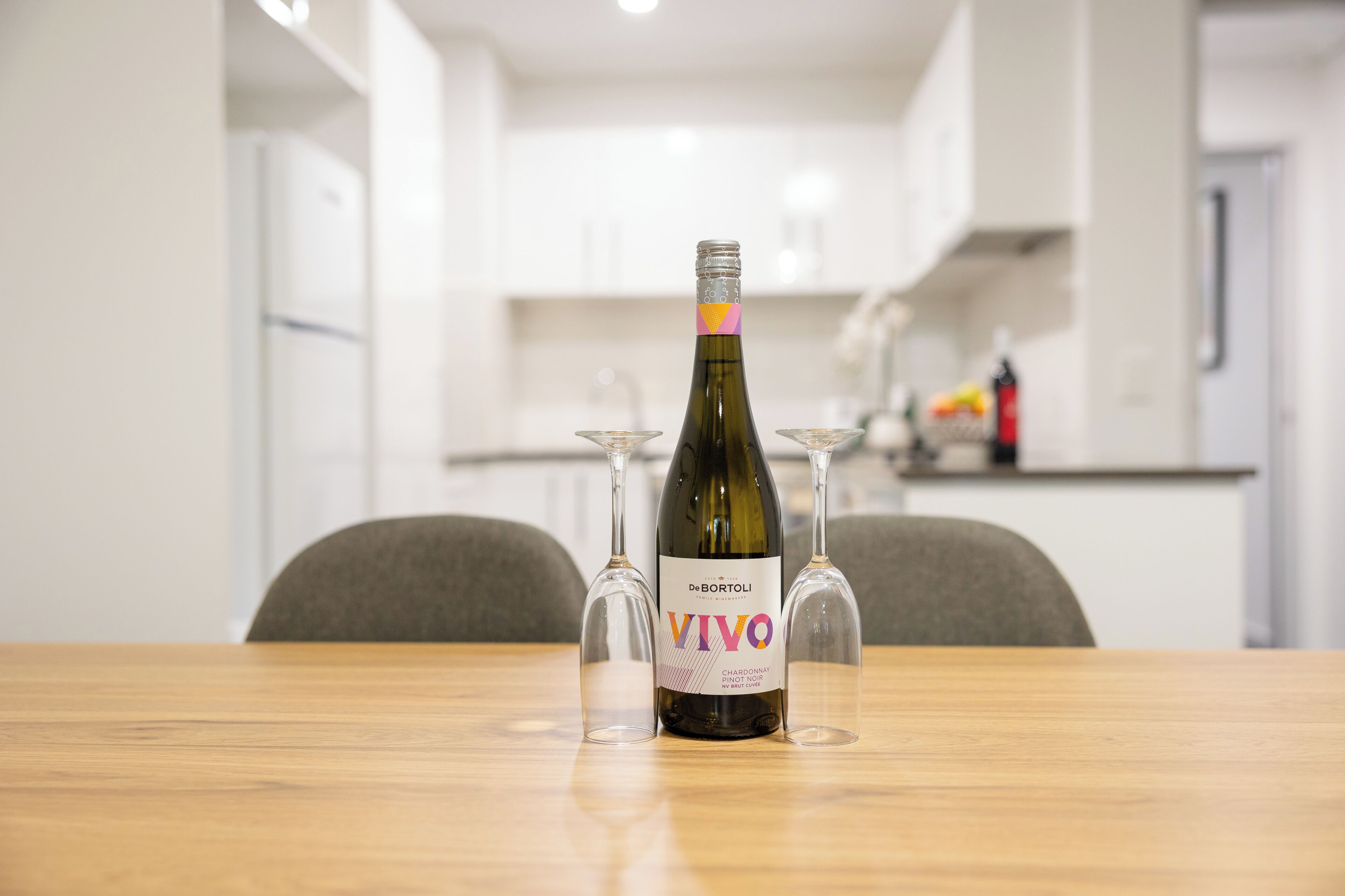 De Bortoli VIVO Chardonnay Pinot Noir -pullo puisella pöydällä, sen vieressä kaksi tyhjää viinilasia.