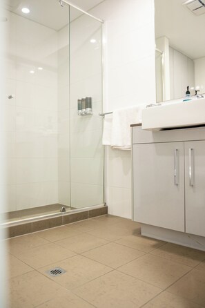 A modern bathroom with a glass shower enclosure, a sink with a mirror, and a cabinet with handles. - Baileys Serviced Apartments (East Perth)