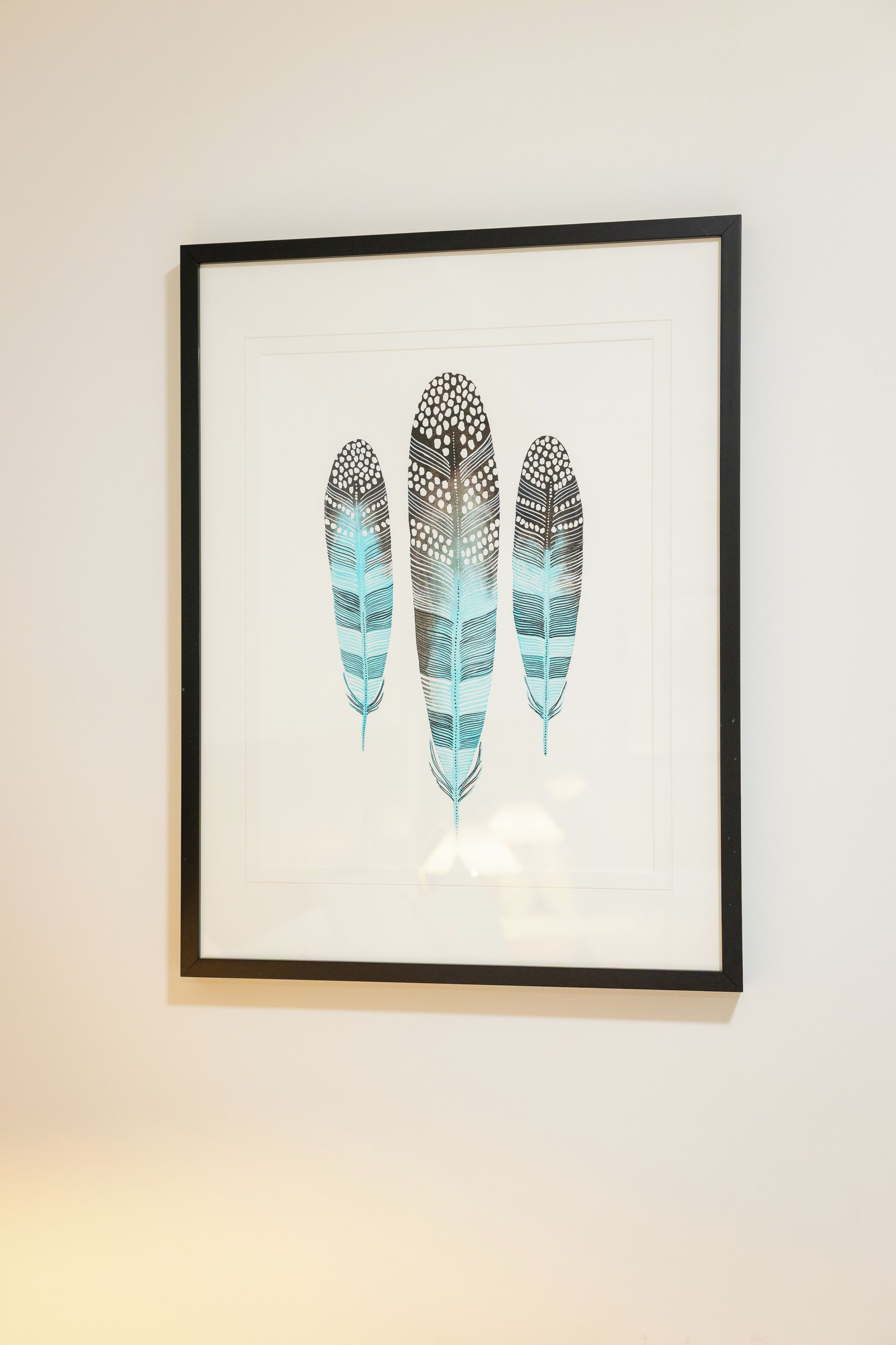 A framed artwork of three stylized feathers on a plain wall.