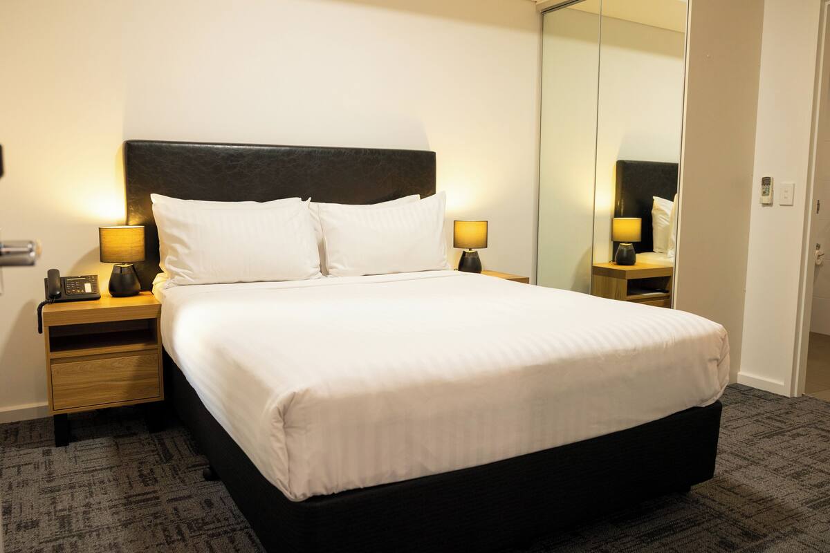 A hotel room with a large bed, two bedside tables with lamps, a nightstand, a mirror, and a carpeted floor.