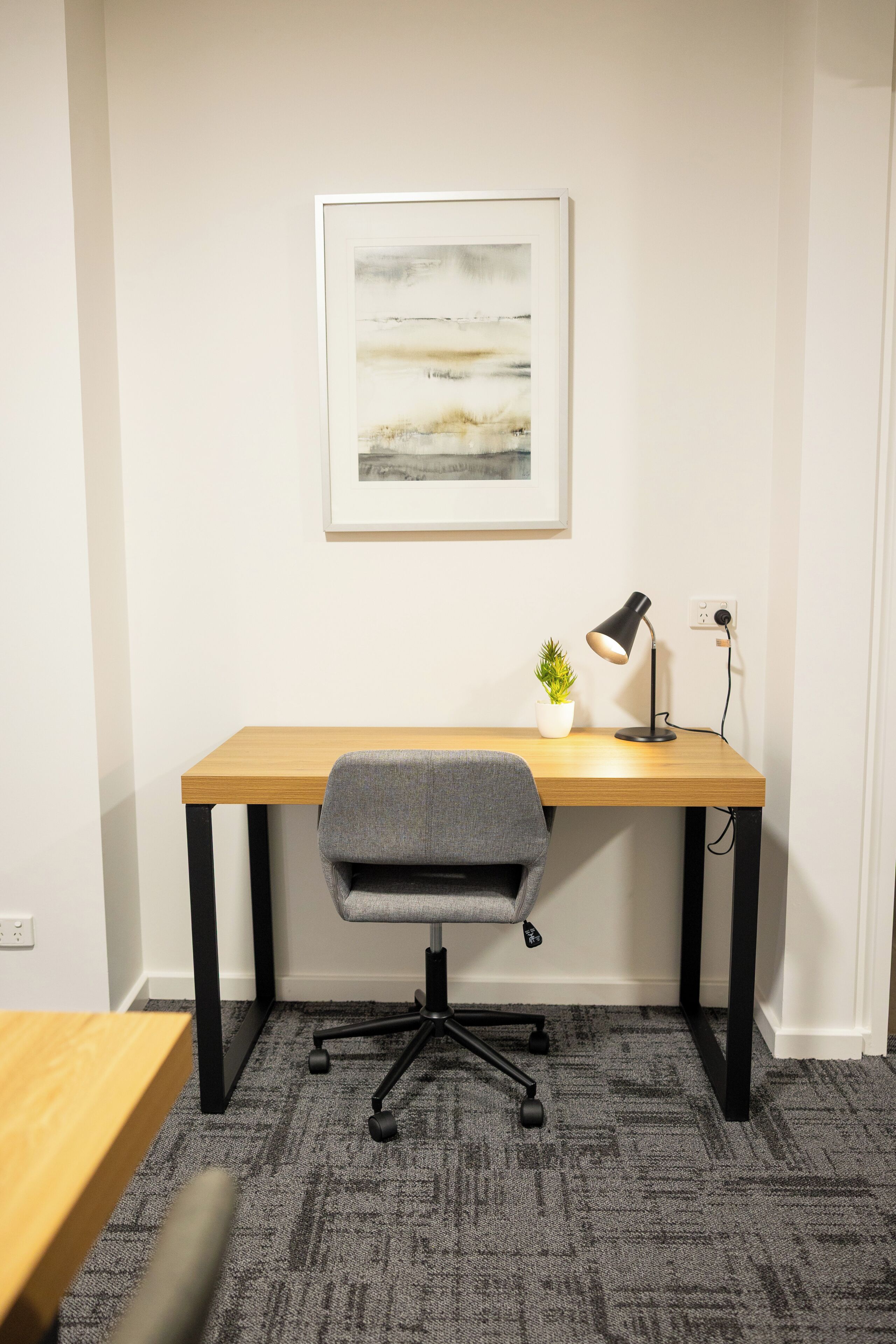 Un espacio de trabajo con escritorio, silla, lámpara y un cuadro en la pared.