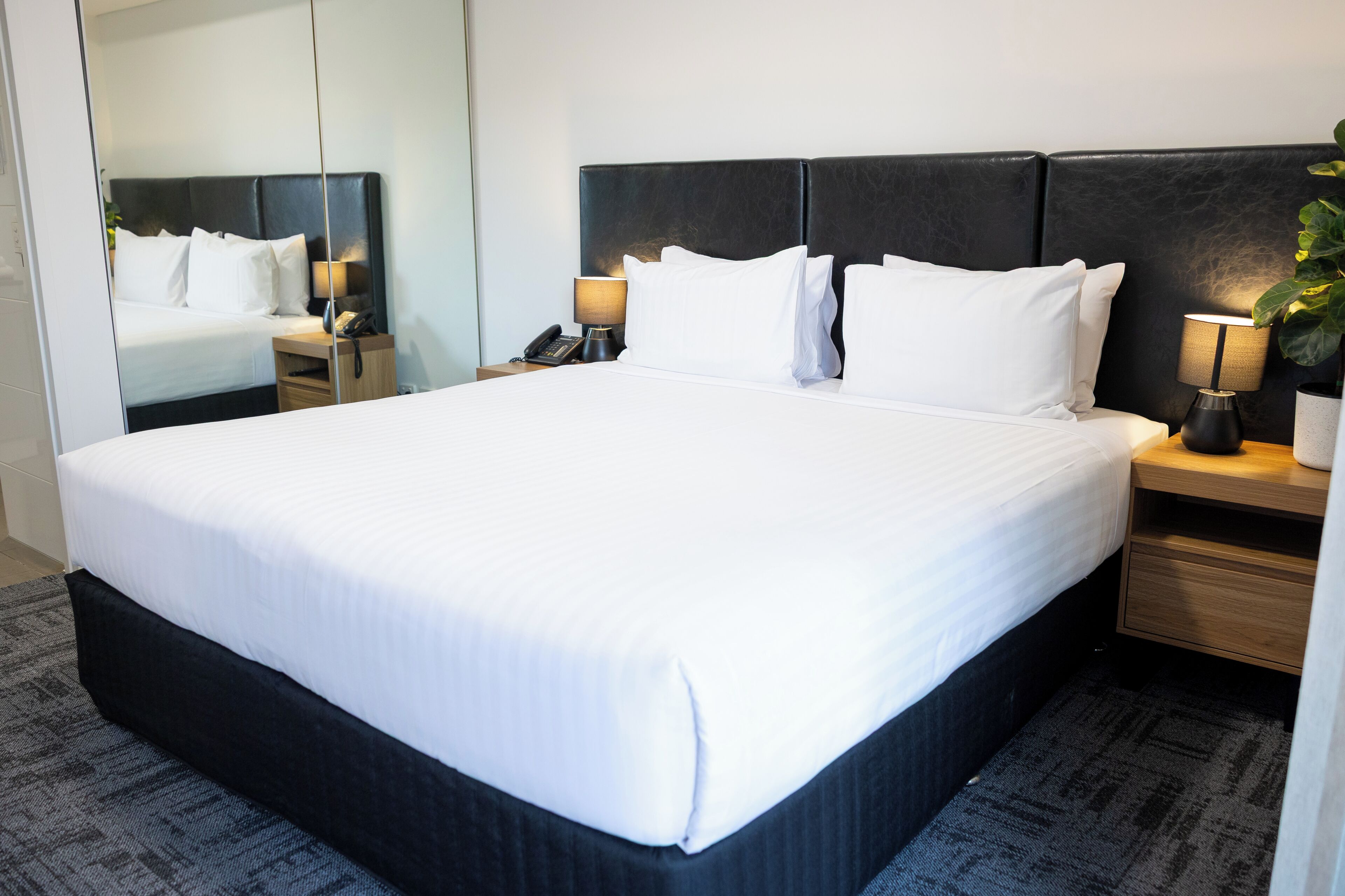 Una habitación de hotel con una cama grande, una mesita de noche con lámpara, teléfono y espejo.