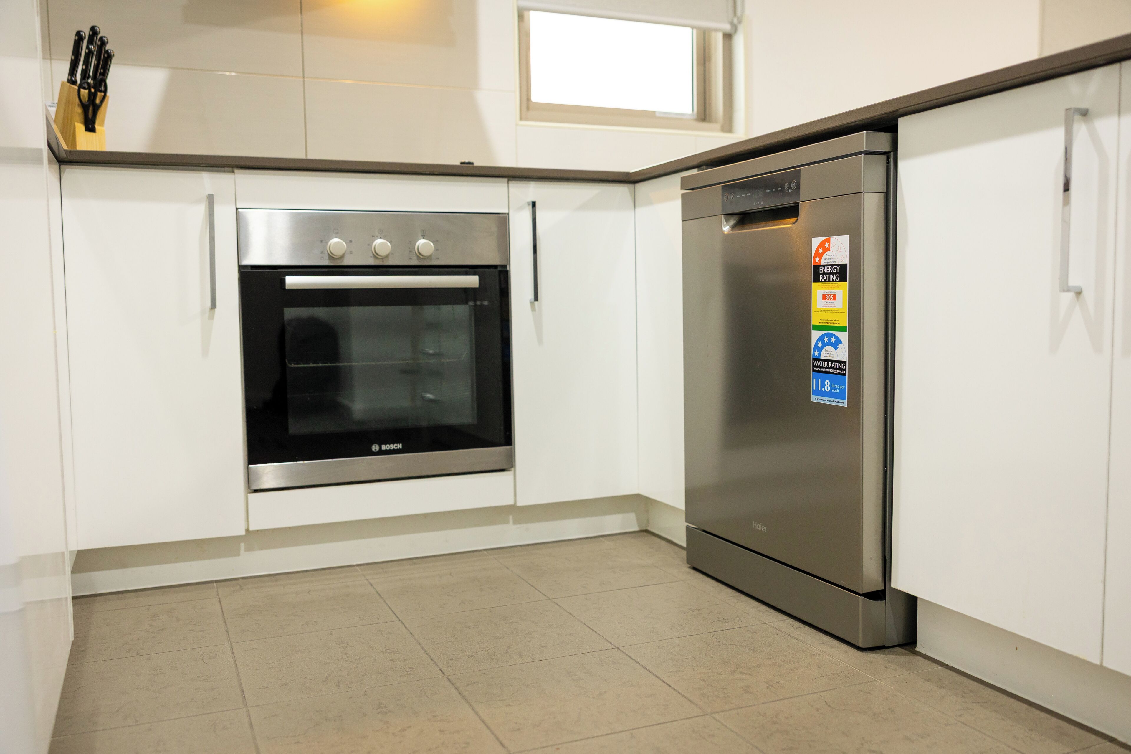 Une cuisine équipée d’un four Bosch et d’un réfrigérateur en acier inoxydable, de meubles blancs et d’une fenêtre située au-dessus du four.