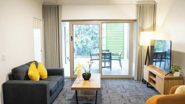 A modern hotel room with a sofa, a small table with a plant, a TV, and a sliding glass door leading to an outdoor area. - Baileys Serviced Apartments (East Perth)