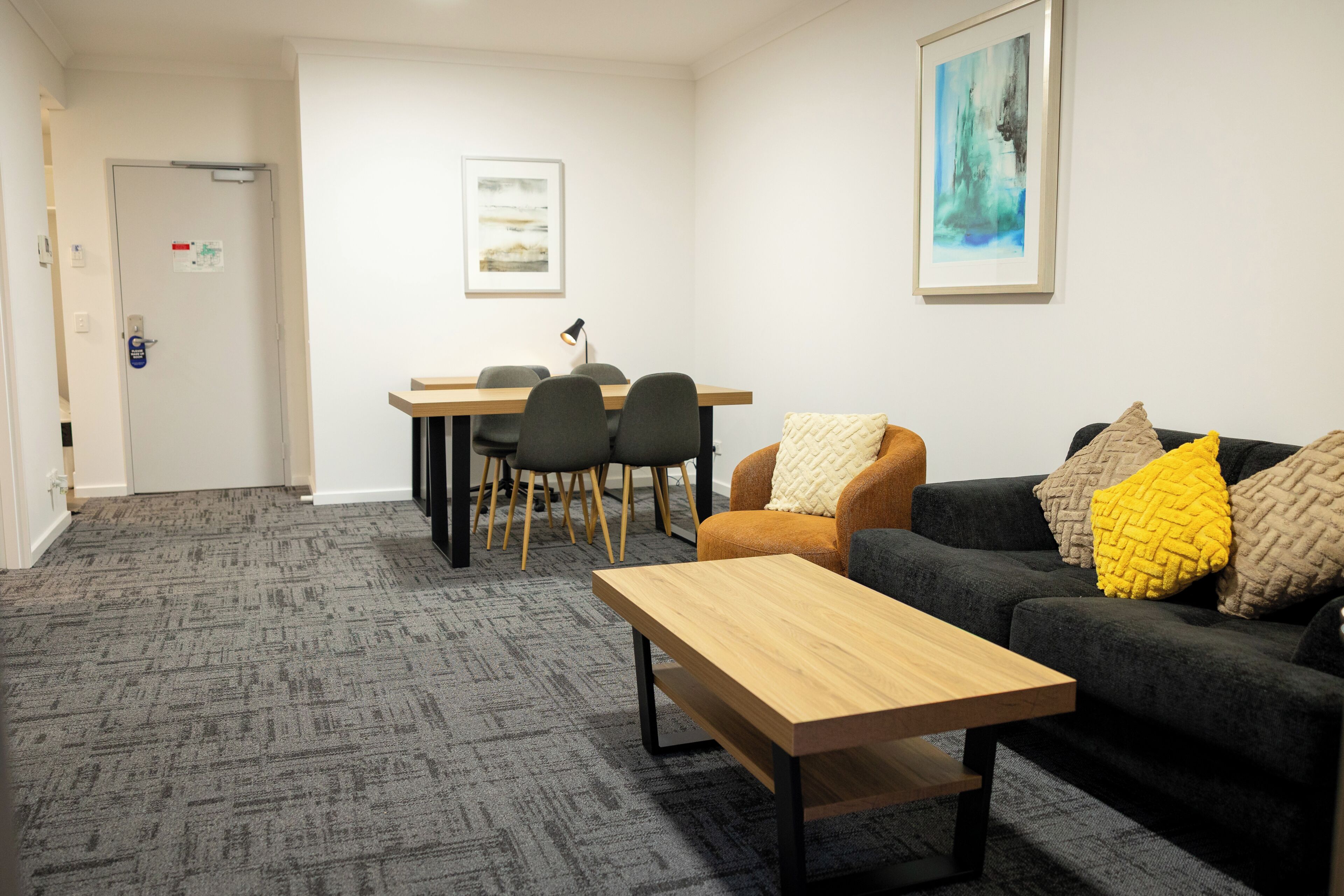 Una habitación con alfombra gris, una mesa de madera, un sofá negro con cojines amarillos y beige, y un comedor con sillas y una mesa.