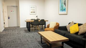 Una habitación con alfombra gris, una mesa de madera, un sofá negro con cojines amarillos y beige, y un comedor con sillas y una mesa.