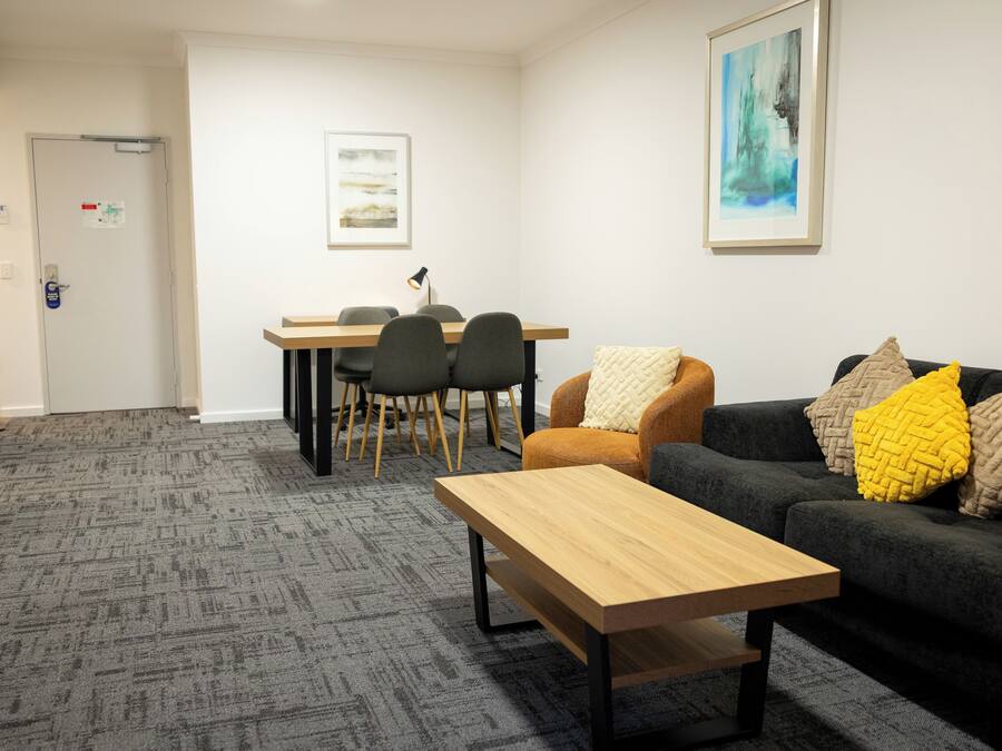 Una habitación con alfombra gris, una mesa de madera, un sofá negro con cojines amarillos y beige, y un comedor con sillas y una mesa.