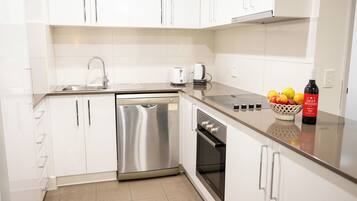 A modern kitchen with white cabinets, a stainless steel dishwasher, an oven, a sink, and a countertop with a fruit bowl and a bottle of wine.