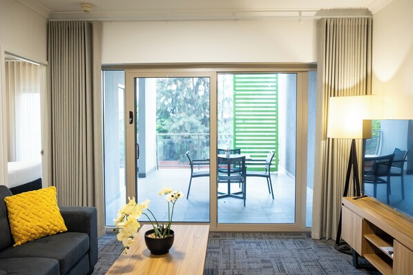 Une chambre d’hôtel avec un canapé, une table basse agrémentée de fleurs, un meuble pour la télévision et une porte-fenêtre coulissante donnant sur une terrasse extérieure avec une table et des chaises.