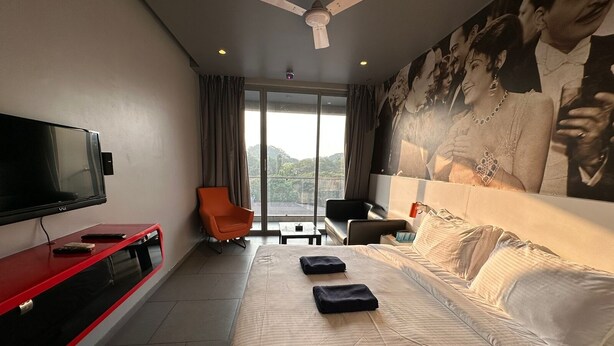 A hotel room with a large bed, a TV, a red console table, an orange chair, and a large window with a view. - SinQ Party Hotel (Candolim)