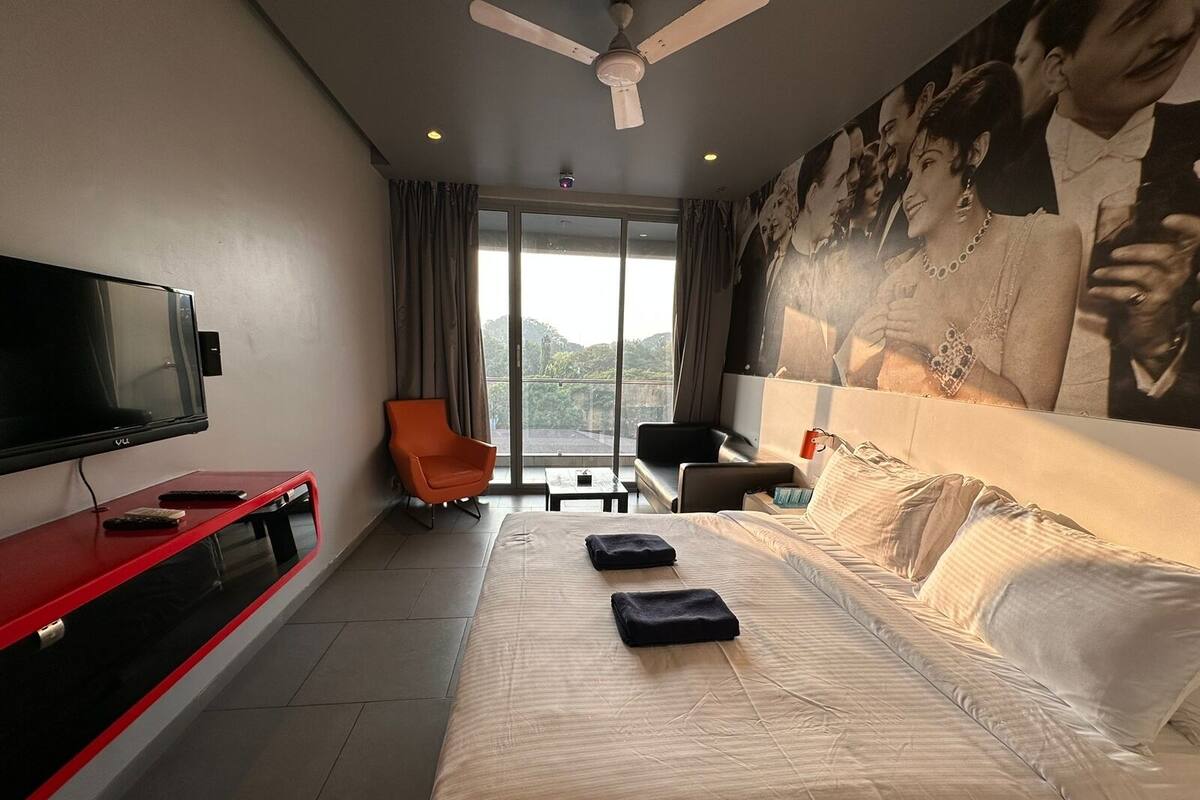 A hotel room with a large bed, a TV, a red console table, an orange chair, and a large window with a view.