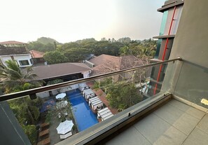 A view from a balcony overlooking a swimming pool with lounge chairs and umbrellas, surrounded by greenery and buildings. - SinQ Party Hotel (Candolim)