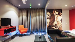 A modern living room with a flat-screen TV, a red entertainment unit, a glass coffee table, a black sofa, an orange chair, and a large wall mural. - SinQ Party Hotel (Candolim)