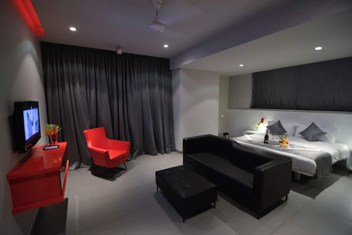 A modern hotel room with a bed, a red chair, a black sofa, a TV, and a desk with a vase of flowers.