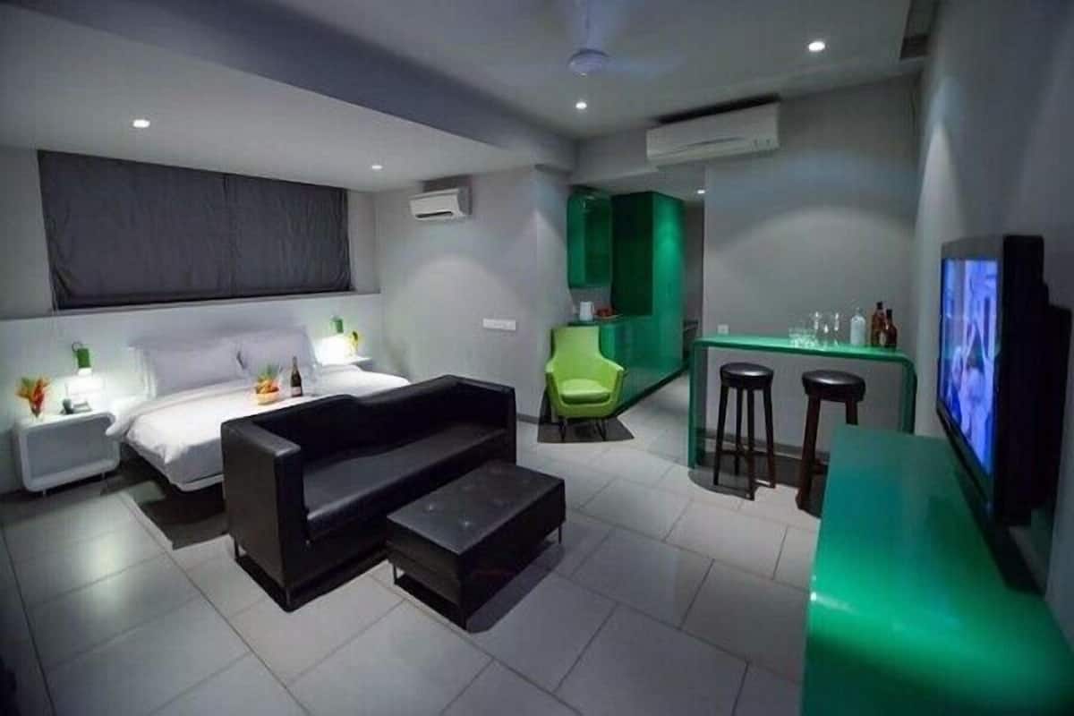 A modern hotel room with a bed, a black sofa, a small coffee table, a green chair, a bar area with stools, and a television.
