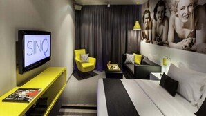 A modern hotel room with a bed, a TV displaying 'SINO', a yellow armchair, a sofa, and a large wall mural. - SinQ Party Hotel (Candolim)