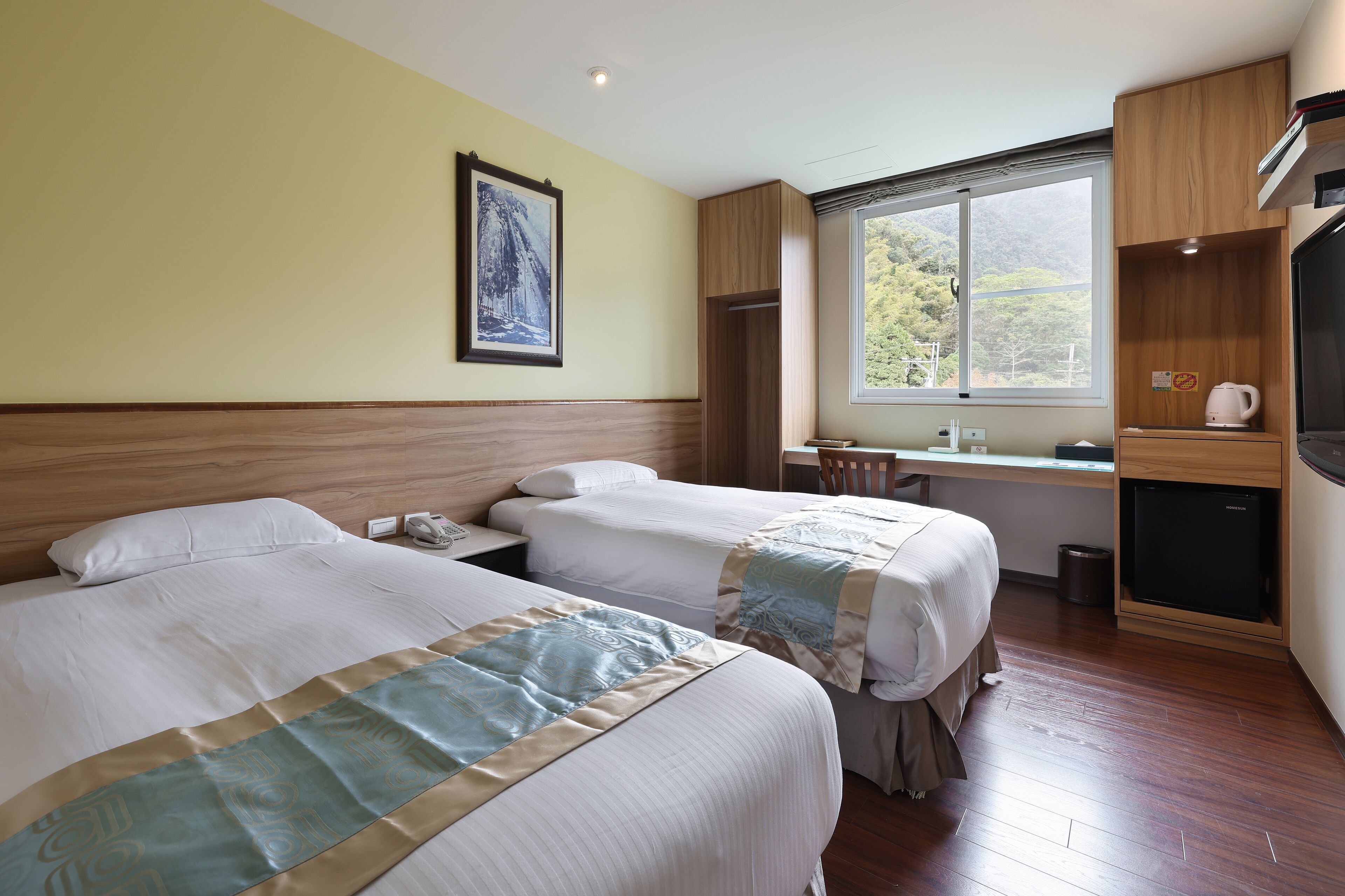 A hotel room with two beds, a desk, a chair, a small refrigerator, and a window with a view of greenery.