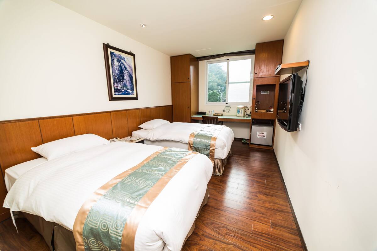 A hotel room with two beds, a desk, a television, and a window with a view of trees.