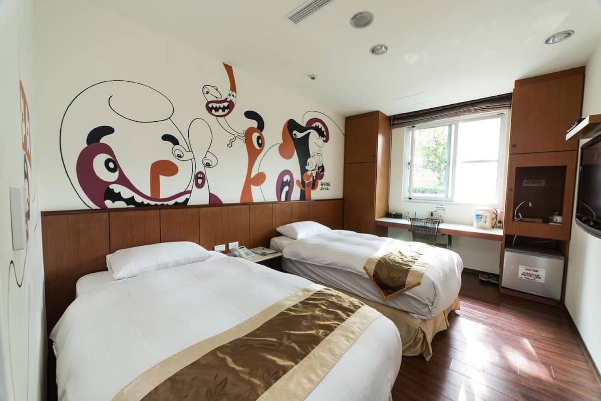 A hotel room with two beds, a desk, a refrigerator, and a wall mural.