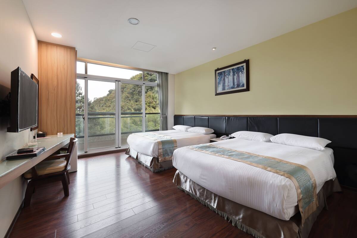 A hotel room with two beds, a desk with a chair, a TV, and a large window offering a view of trees.