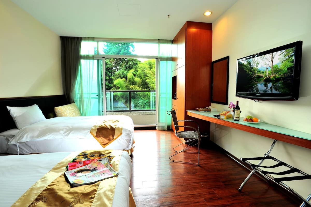 A hotel room with two beds, a desk with a chair, a TV, and a window with a view of greenery.