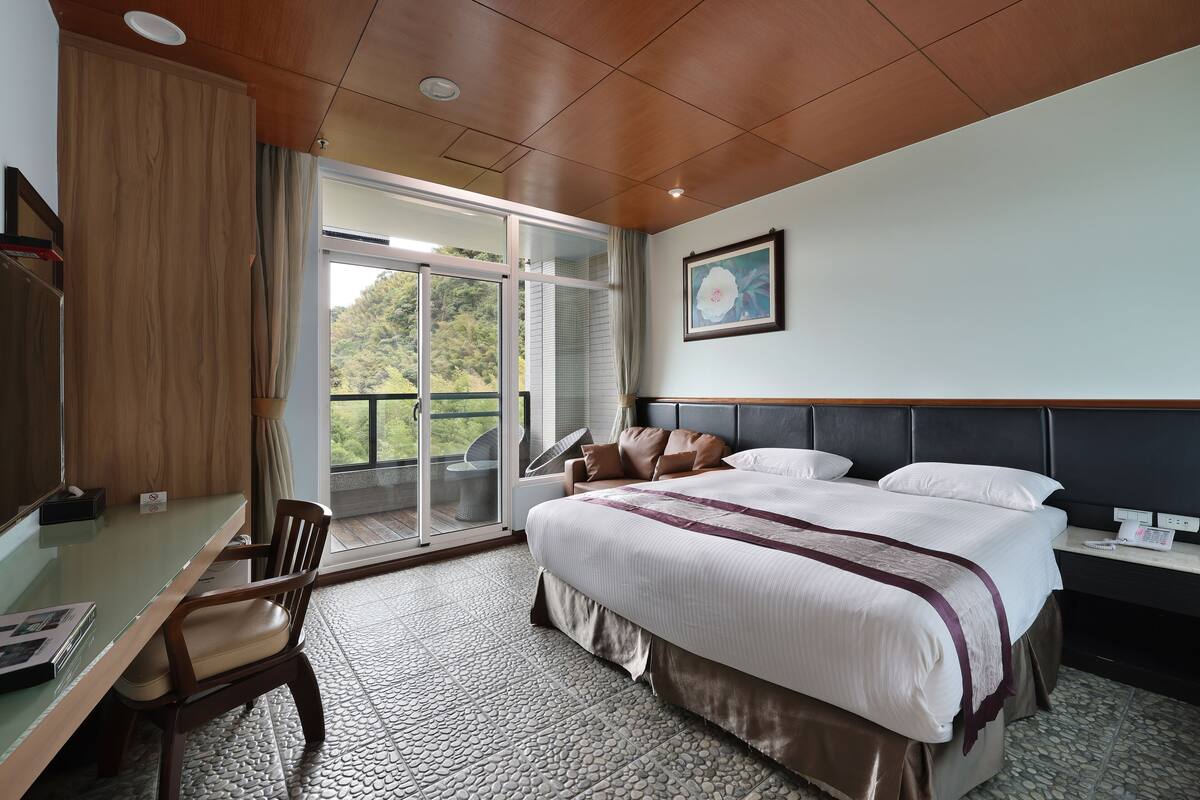 A hotel room with a bed, a desk, a chair, a large window, and a balcony.