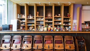 Uma área de bar com prateleiras de madeira exibindo diversos frascos e copos, e várias cadeiras de madeira dispostas na frente.