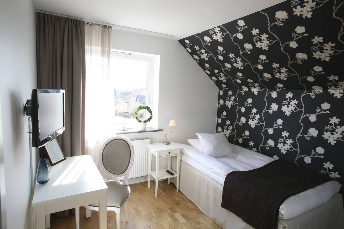 A bedroom with a bed, a desk with a chair, a TV, and a window with curtains.