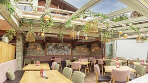 A restaurant with multiple wooden tables and chairs, hanging plants, and a stone wall with a floral painting. - Hotel San Marco (Fuessen)